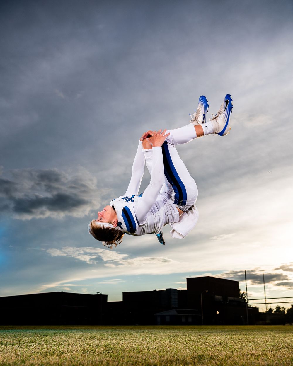 Athletic person in white uniform performs a dynamic backflip against a dramatic cloudy sky over grassy field at sunset.