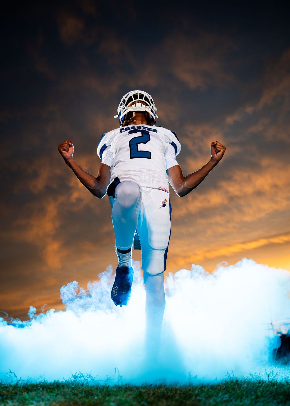 Football player wearing jersey number 2 strikes dramatic pose with smoke effects against vibrant sunset sky.