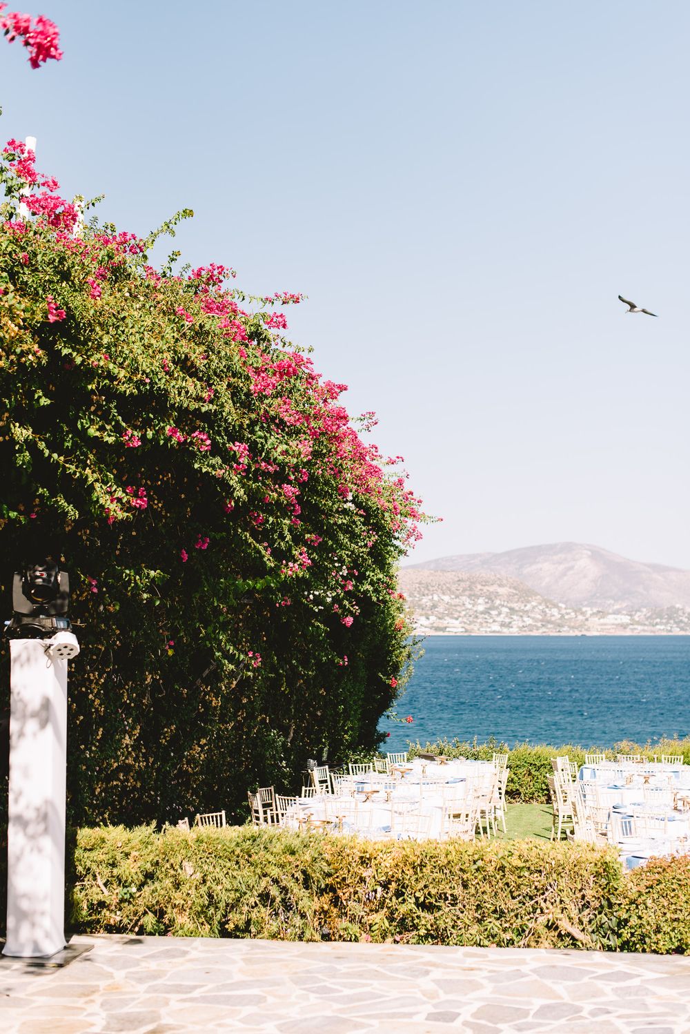 Outdoor wedding venue with pink bougainvillea, white chairs, and stunning sea and mountain views under clear blue sky.