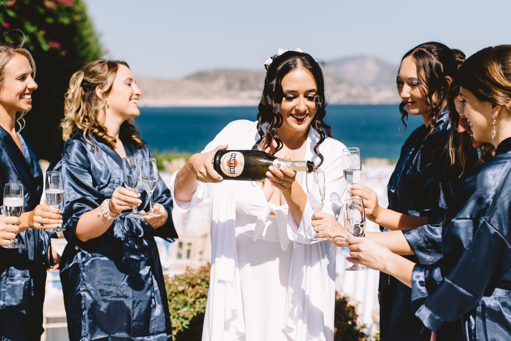 Bride in white robe pouring champagne for bridesmaids in navy satin robes outdoors by the ocean.