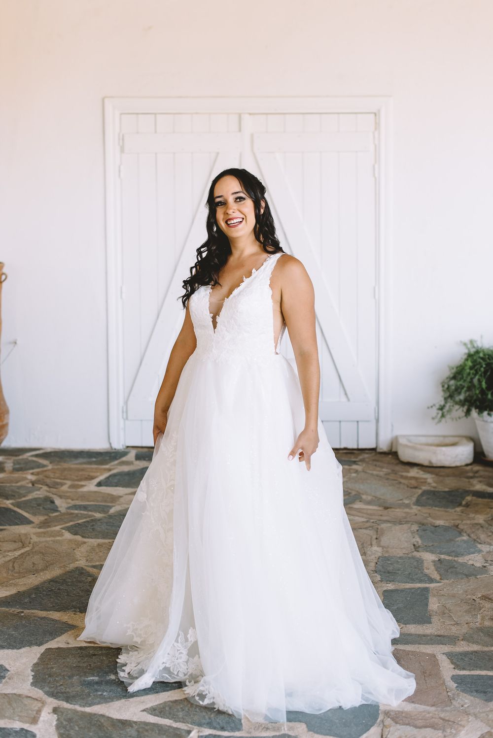 Smiling bride in a white lace A-line wedding gown with veil, standing on stone patio before white doors.