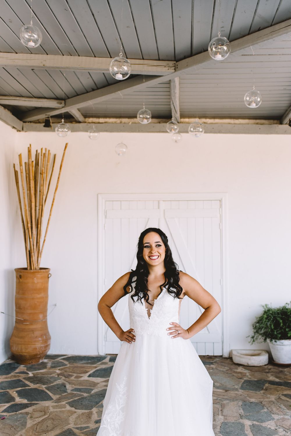 Smiling bride in white lace wedding gown with veil, standing hands on hips in front of white door on stone patio.