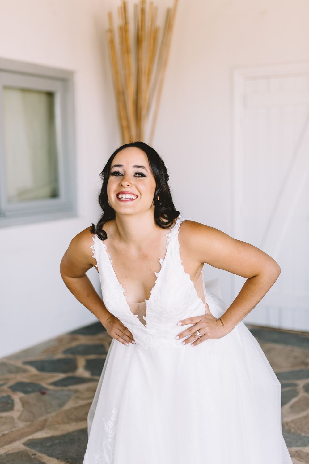 Smiling bride in white lace V-neck wedding gown posing with hands on hips in a bright white room.