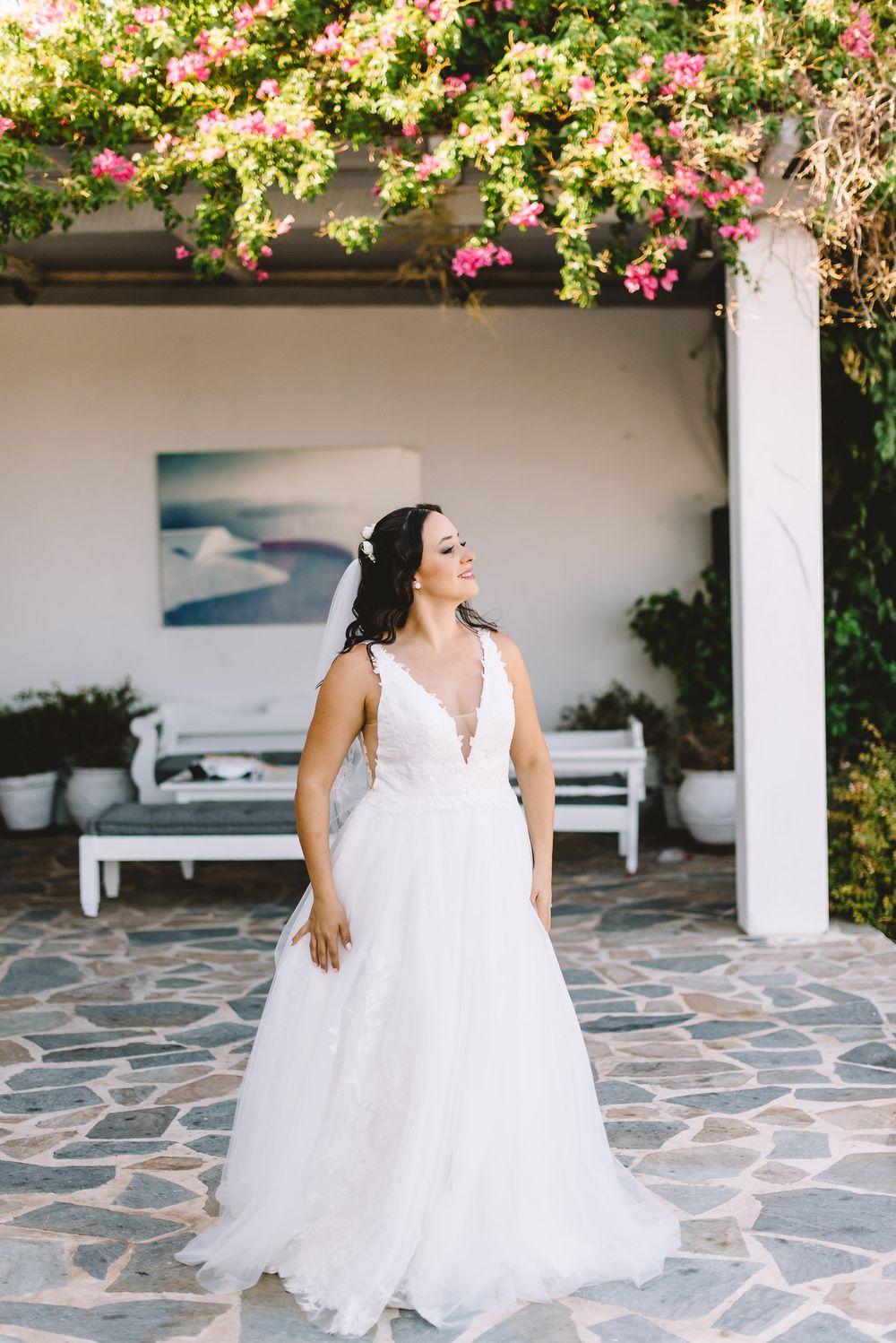 Bride in white lace V-neck wedding gown stands on stone patio beneath blooming pink bougainvillea.