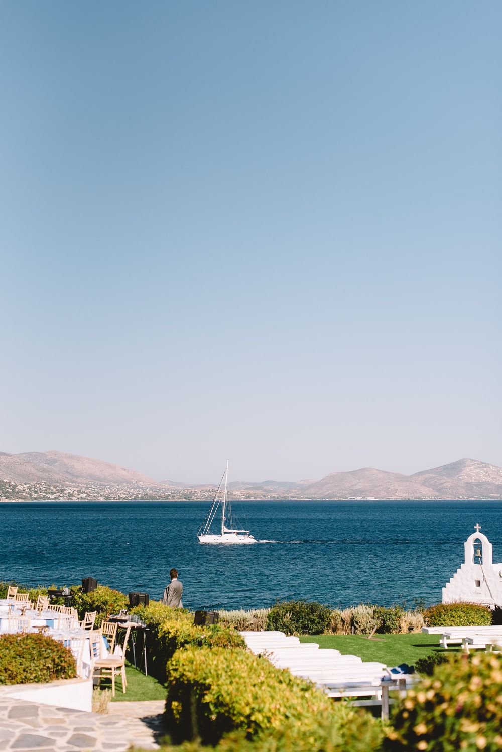 Scenic Greek seaside venue with white chapel, sailboat on blue Aegean Sea, and mountain backdrop under clear sky.