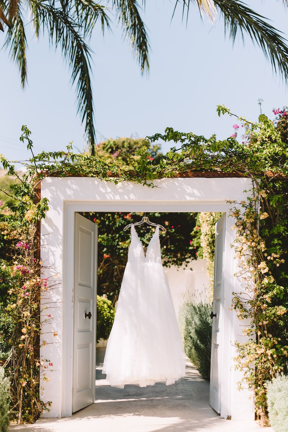 White wedding dress hanging in a floral archway with palm trees and blue sky in a Mediterranean garden.