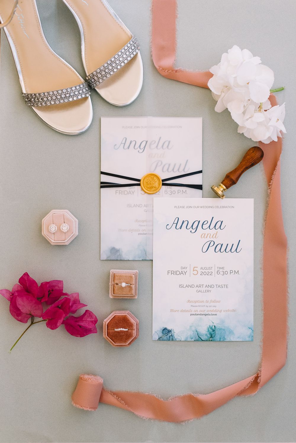 Elegant wedding flat lay with Angela and Paul invitations, diamond sandals, gold wax seal, rings in velvet boxes, and pink ribbons.
