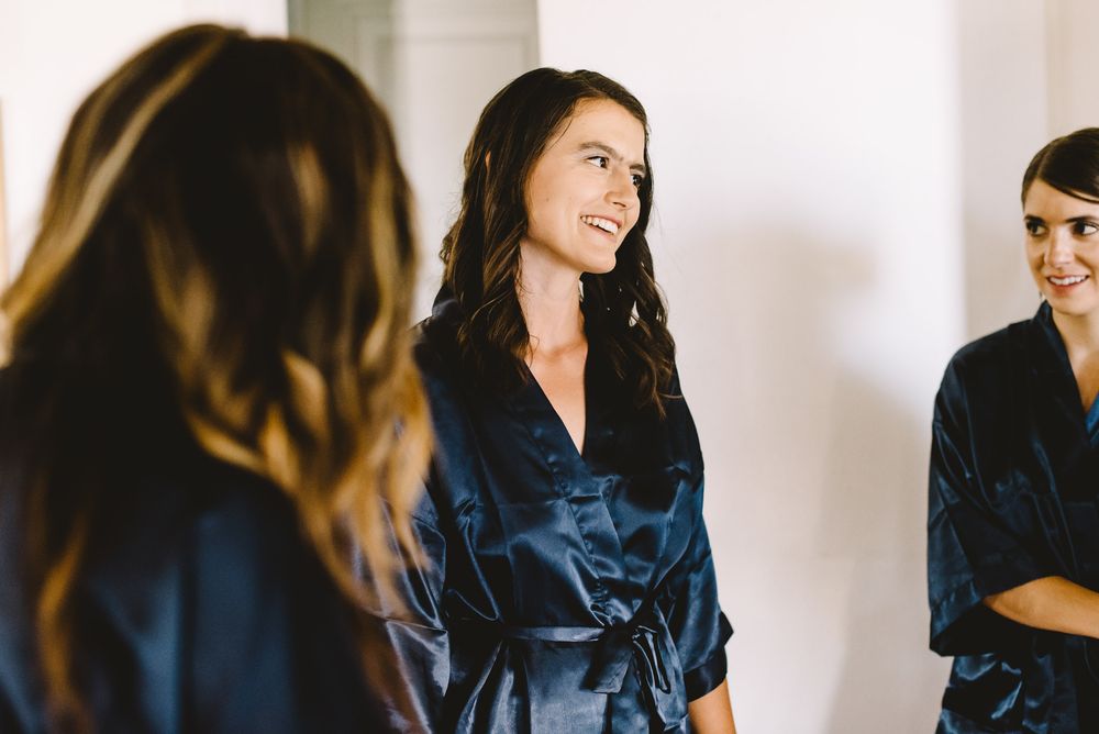Two bridesmaids in navy satin robes smiling and chatting during wedding preparations.