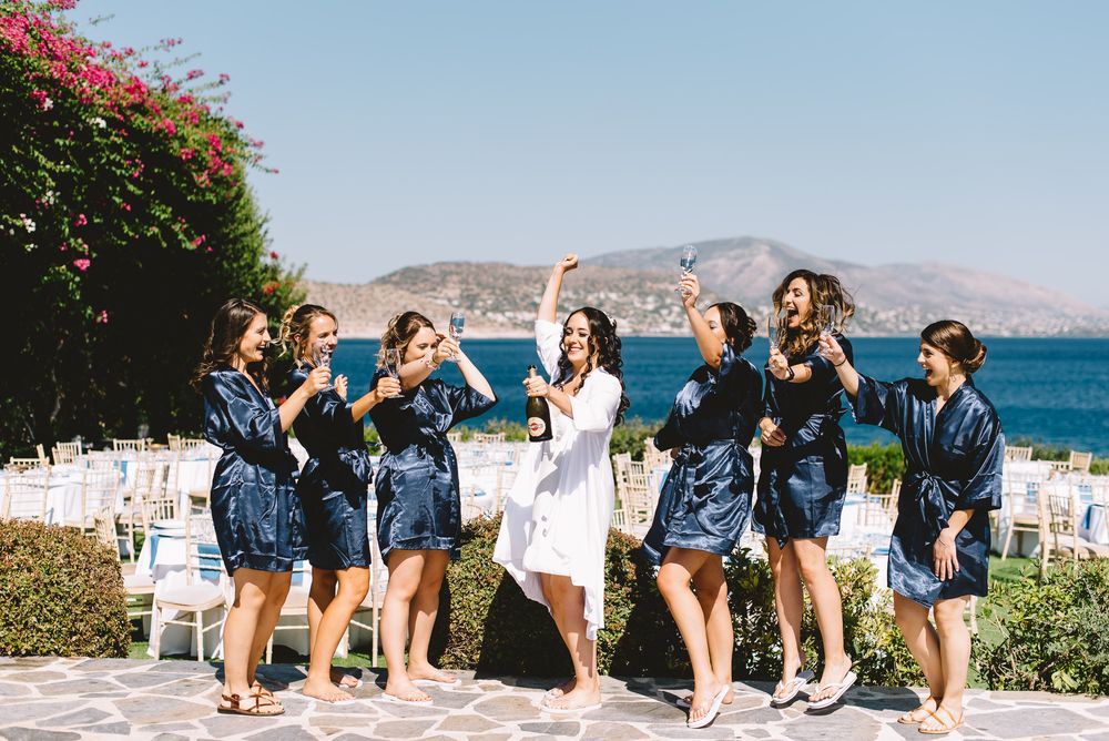 Bridesmaids in navy satin robes celebrate with bride outdoors at a Greek seaside wedding venue.