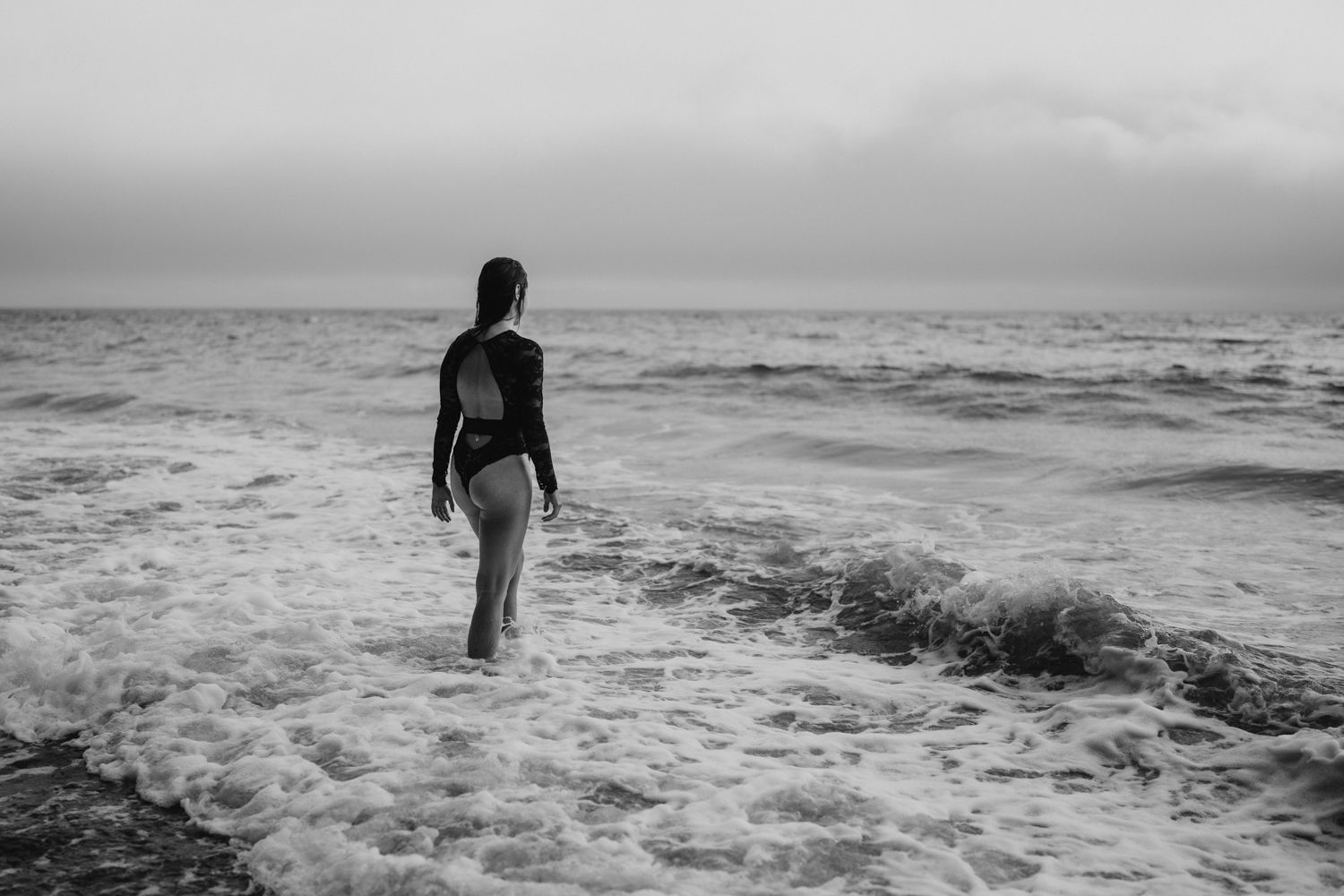 Black and white photo of woman in bikini standing in ocean waves on overcast day.