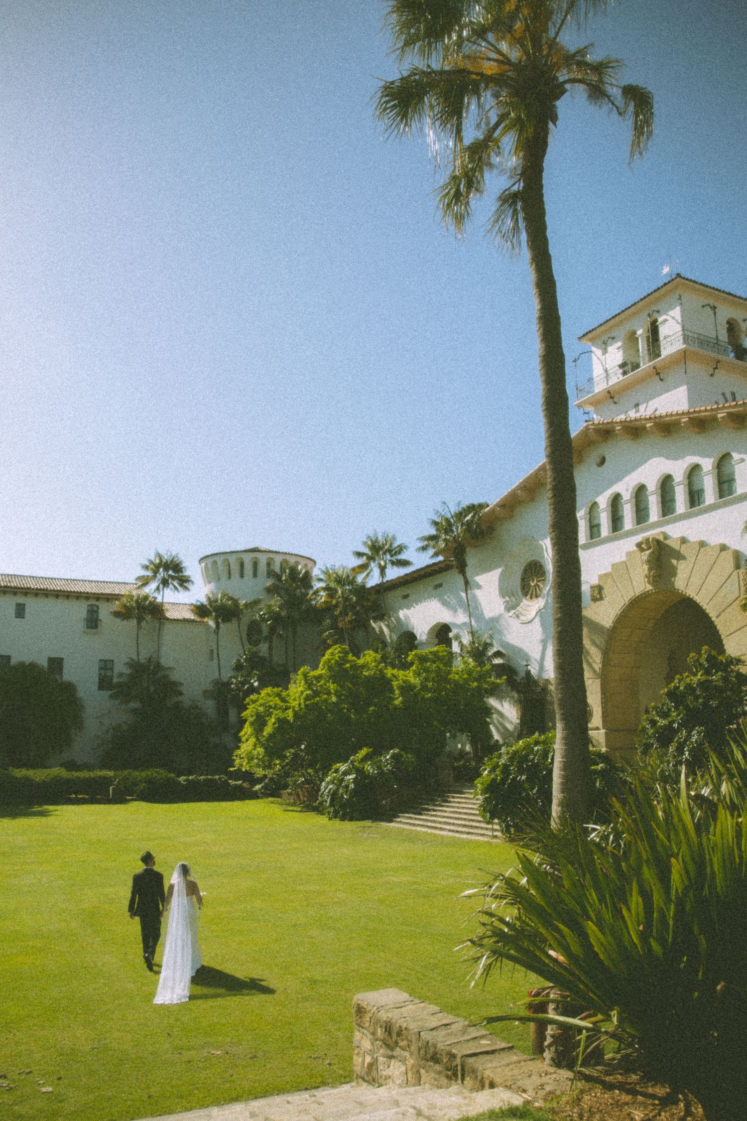 Bride and groom walking on green lawn at Santa Barbara Courthouse wedding venue with palm trees and blue sky.