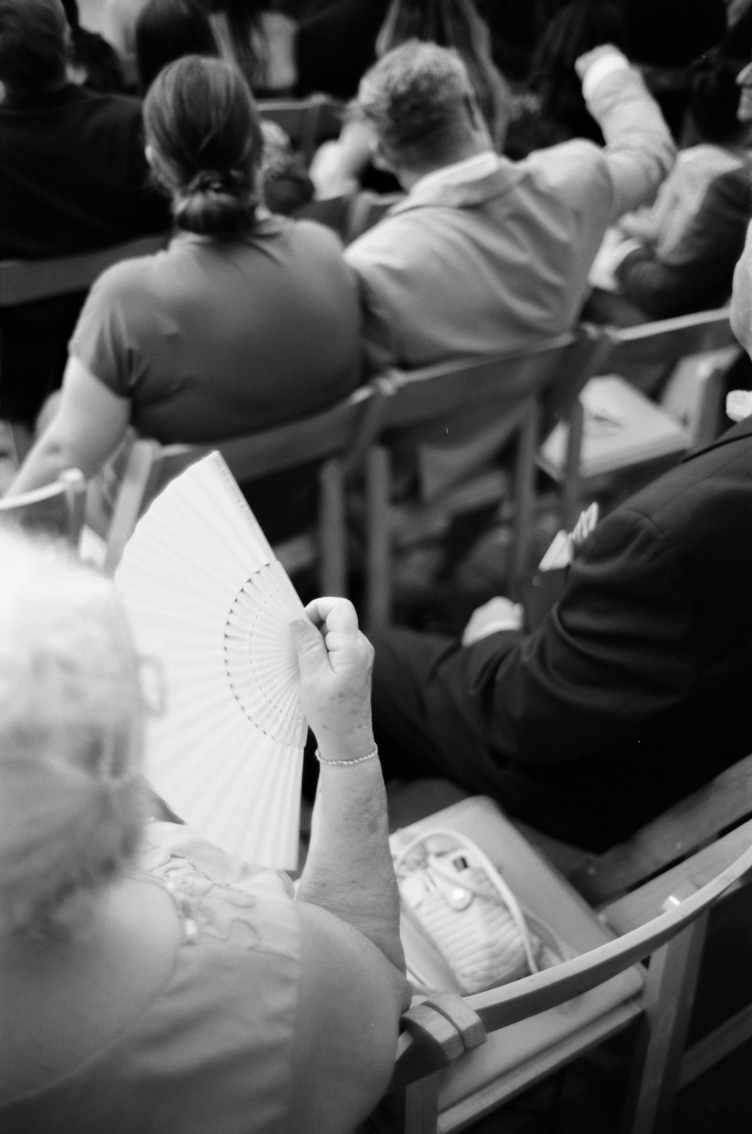 Black and white analog photo of audience members seated in chairs, with a woman holding an open hand fan in the foreground.