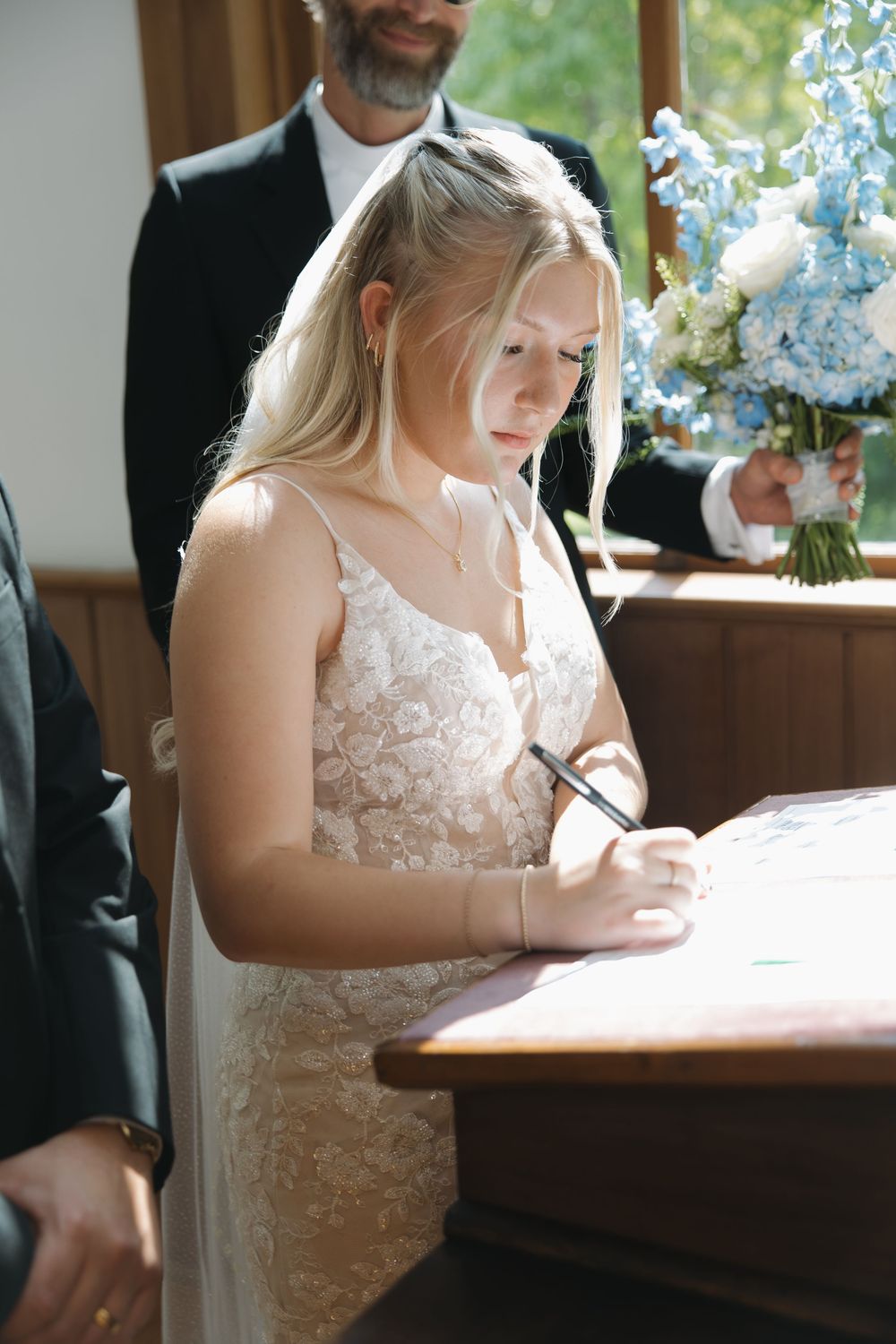 Bride in lace wedding dress signing marriage certificate at wooden table during wedding ceremony.