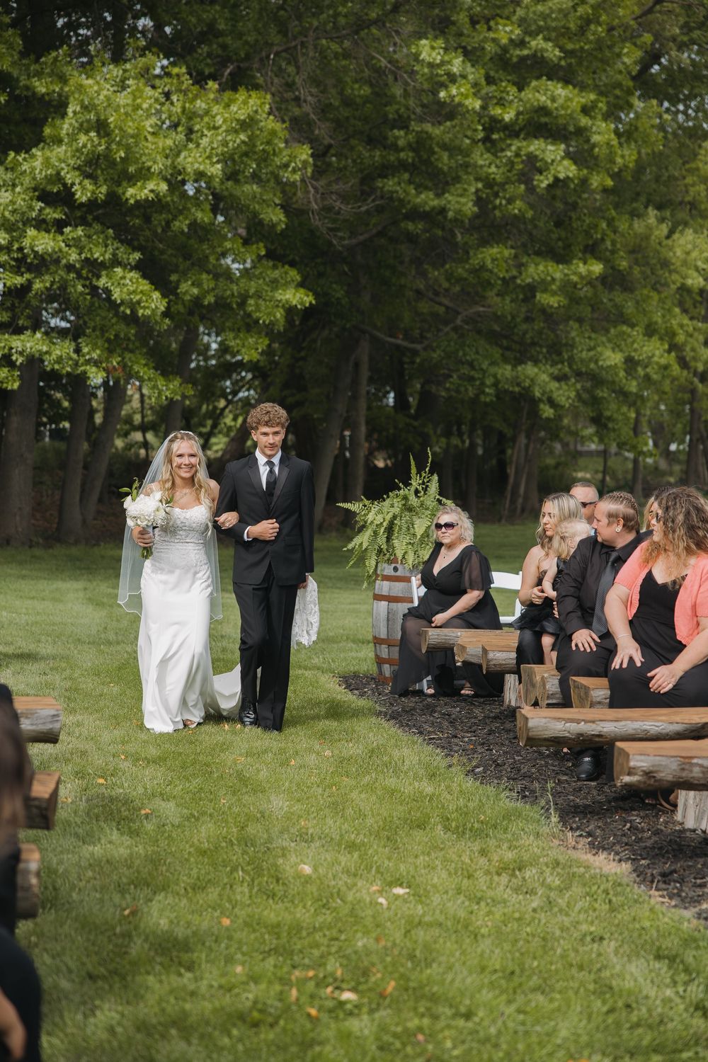 Bride in white gown and veil walks down outdoor aisle with escort past guests seated on wooden benches.