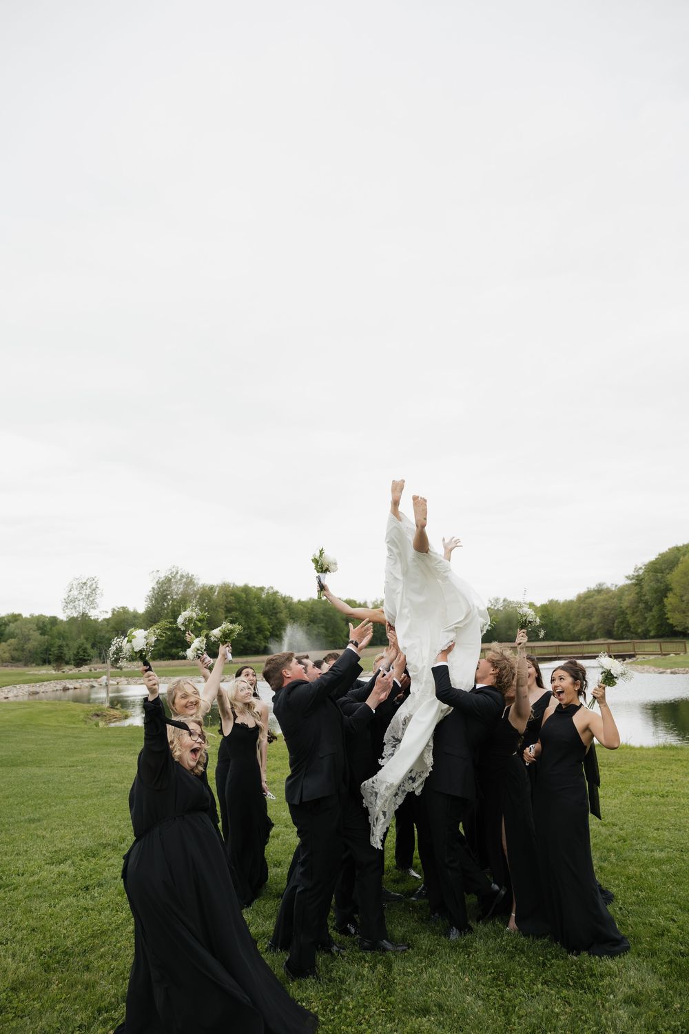 Wedding party tosses bride in white gown into the air outdoors near a pond in celebration.
