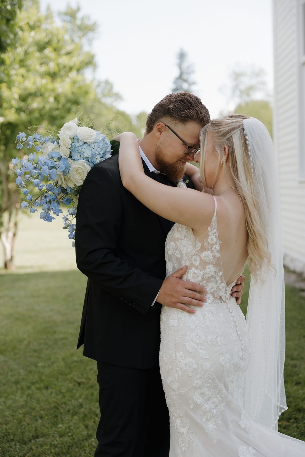 Bride and groom share a romantic forehead touch outdoors, she holds blue and white bouquet, wearing lace gown and veil.