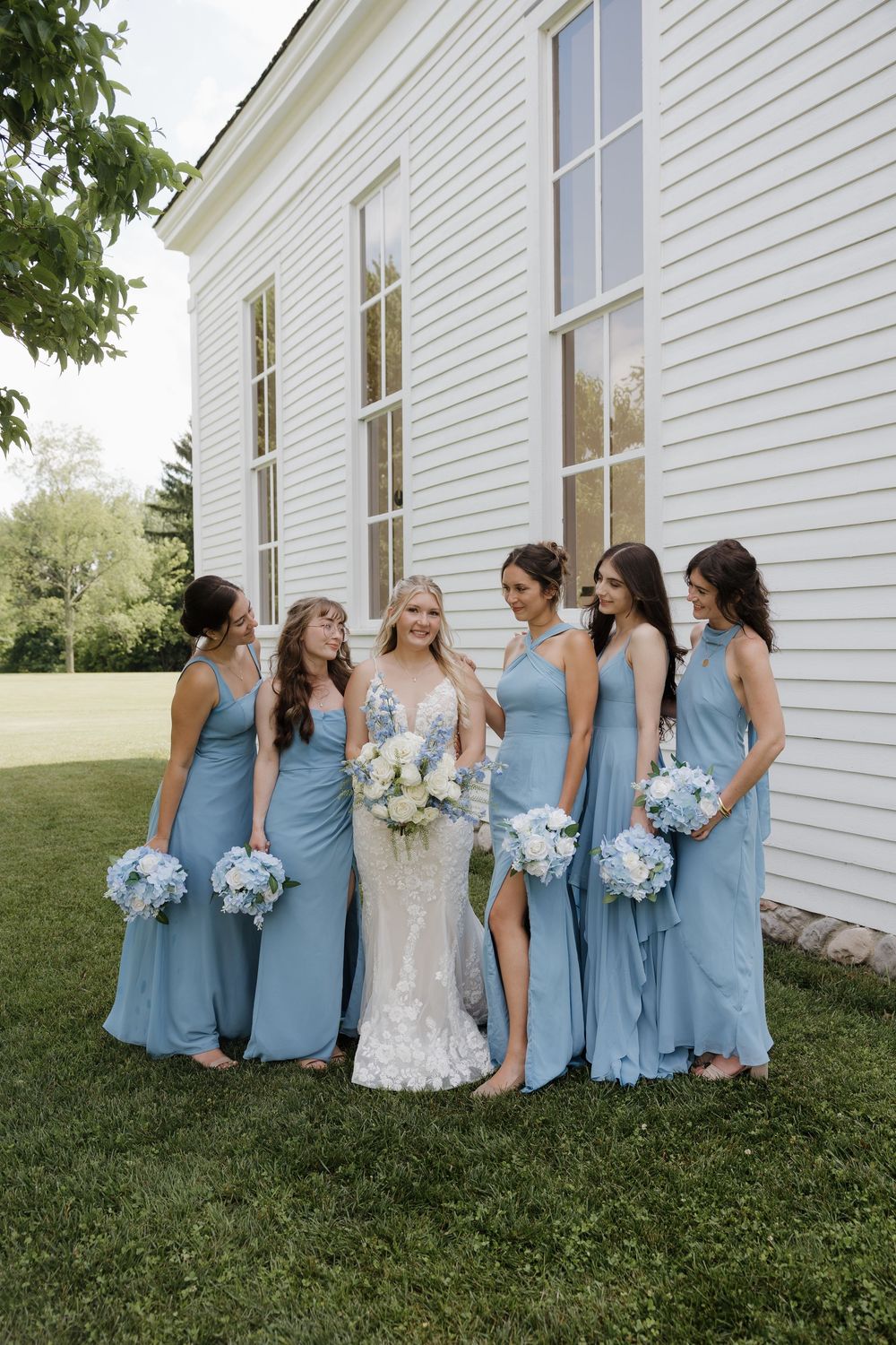 Bride in lace gown with five bridesmaids in dusty blue dresses holding white bouquets outside white building.