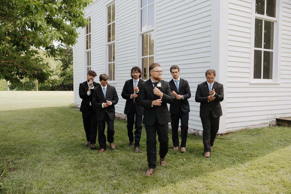 Groom and groomsmen in black suits adjusting bowties outside a white wooden chapel on a sunny day.