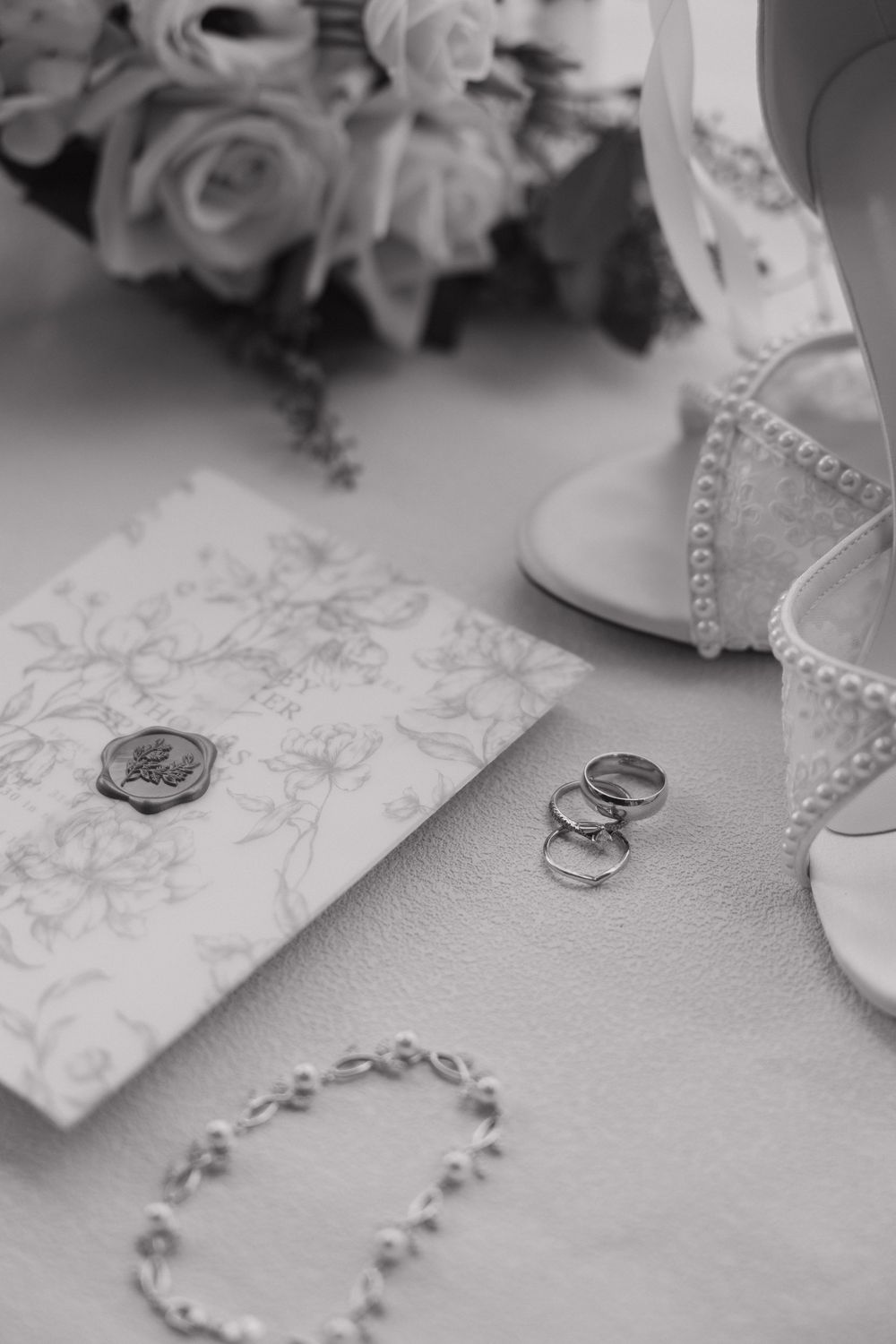 Black and white flat lay of wedding rings, floral envelope, pearl bracelet, bridal heels, and rose bouquet.