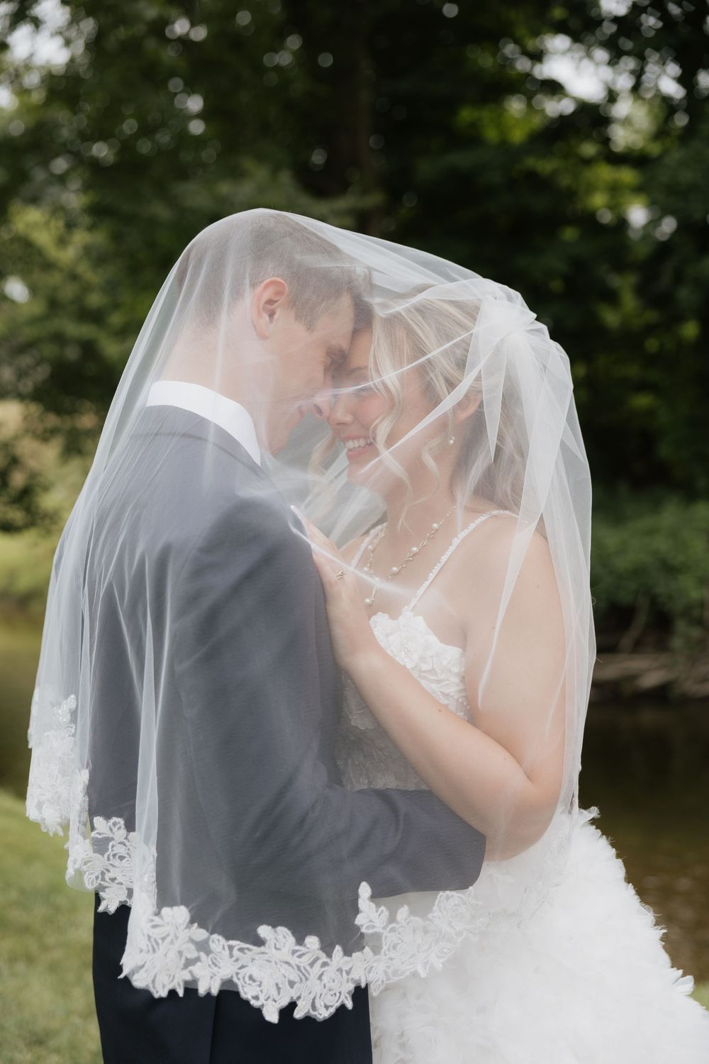 Bride and groom share an intimate moment under a lace-trimmed wedding veil outdoors near a pond.