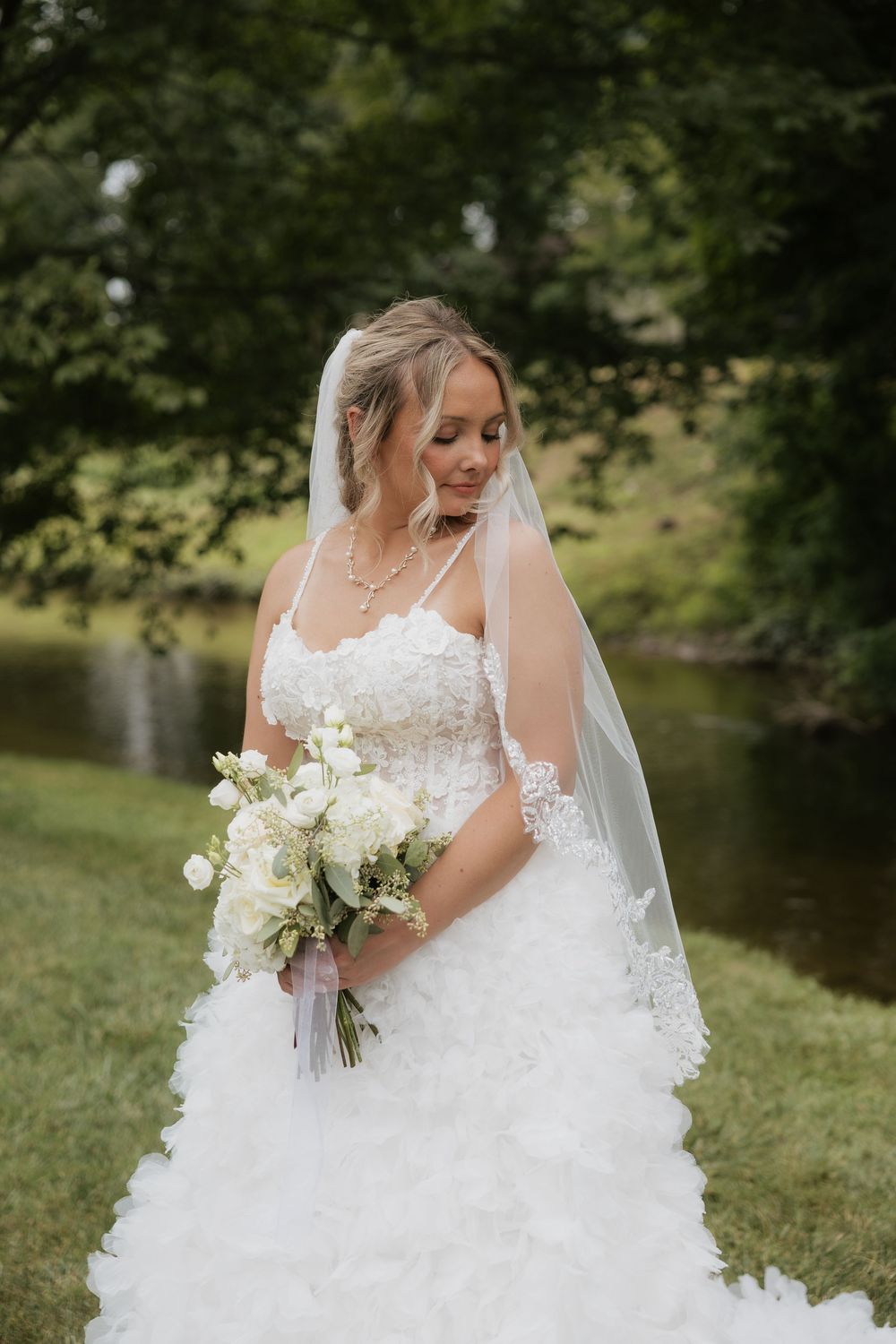 Bride in lace strapless gown and veil holds white bouquet outdoors near pond with lush green trees.