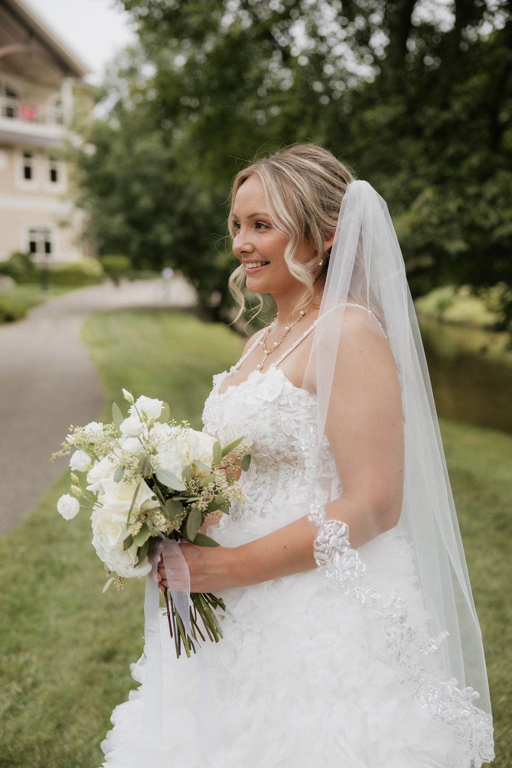 Smiling bride in white lace gown and veil holds white floral bouquet outdoors on her wedding day.