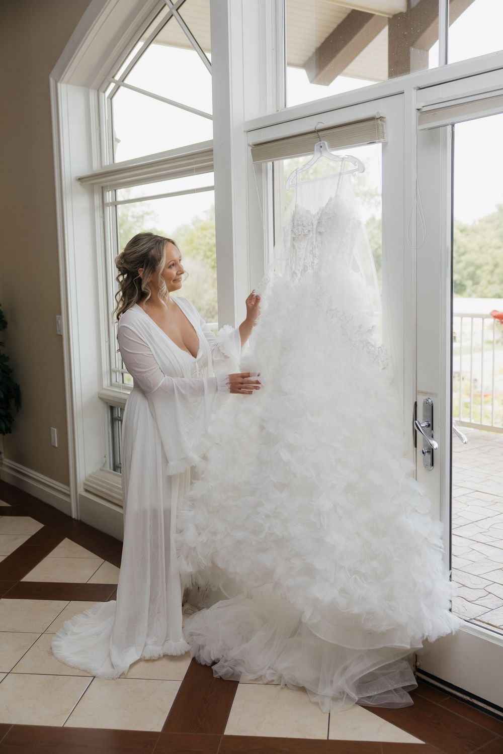 Bride in white robe admiring her fluffy layered wedding gown hanging by large windows before ceremony.