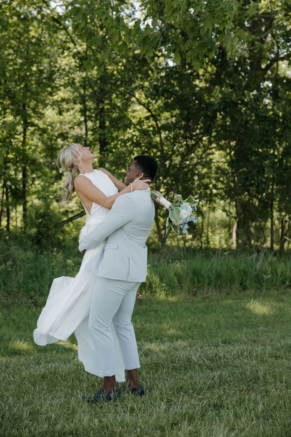 Two brides in white celebrate their wedding day outdoors, one lifting the other in a lush green garden setting.