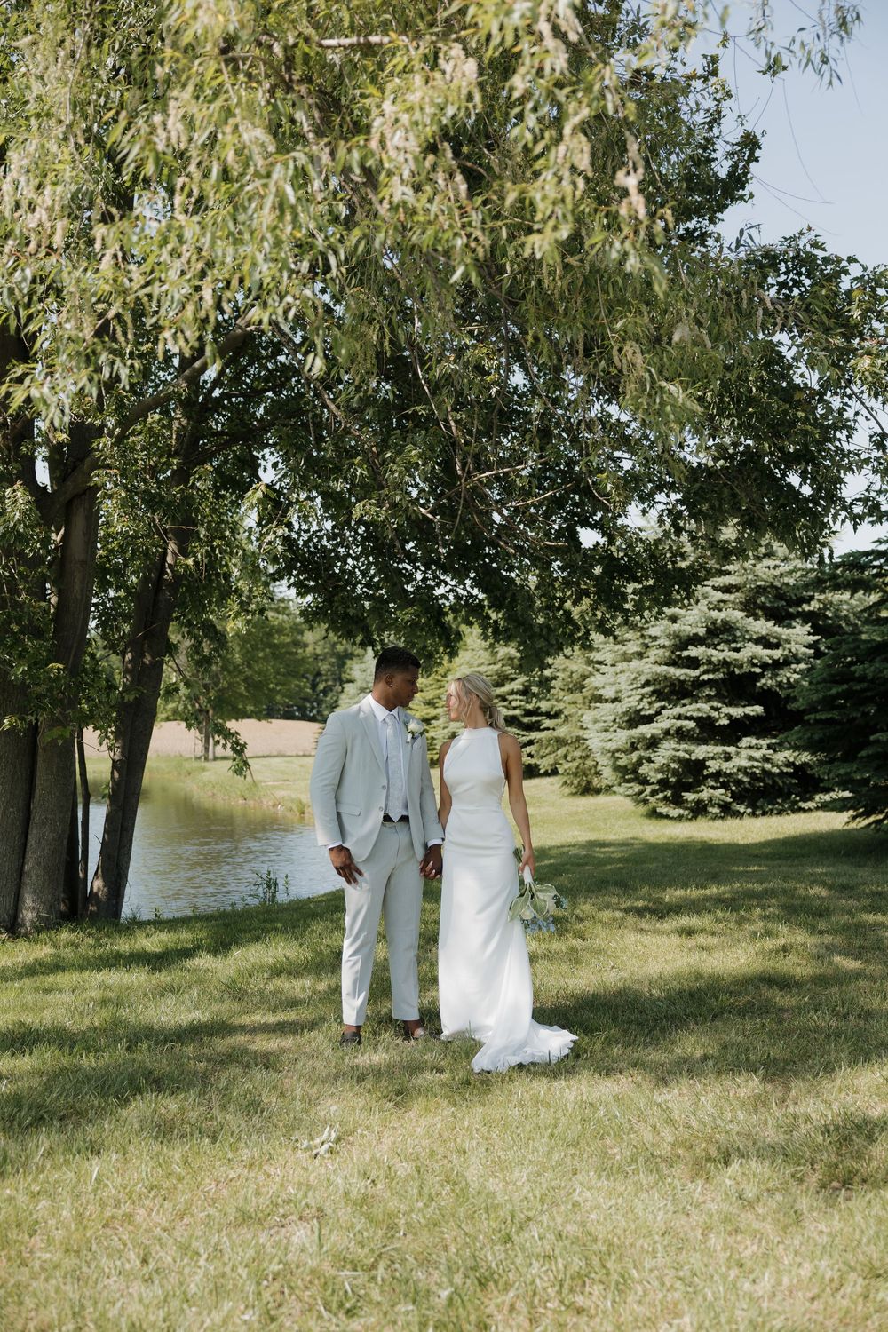 A bride and groom stand hand in hand on a lush green lawn beneath a willow tree near a serene pond on their wedding day.
