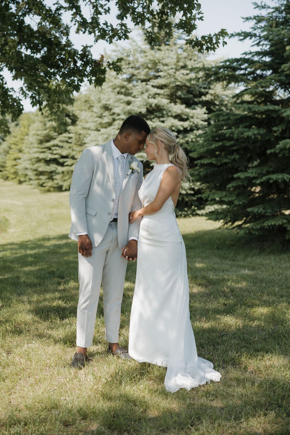 Interracial couple sharing a romantic forehead touch outdoors, groom in light gray suit, bride in white gown, surrounded by evergreen trees.
