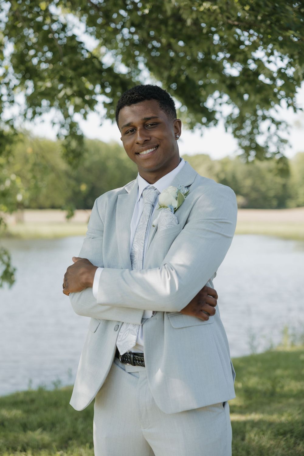Smiling groom in light gray suit with white boutonniere stands arms crossed by a serene lake in a park.