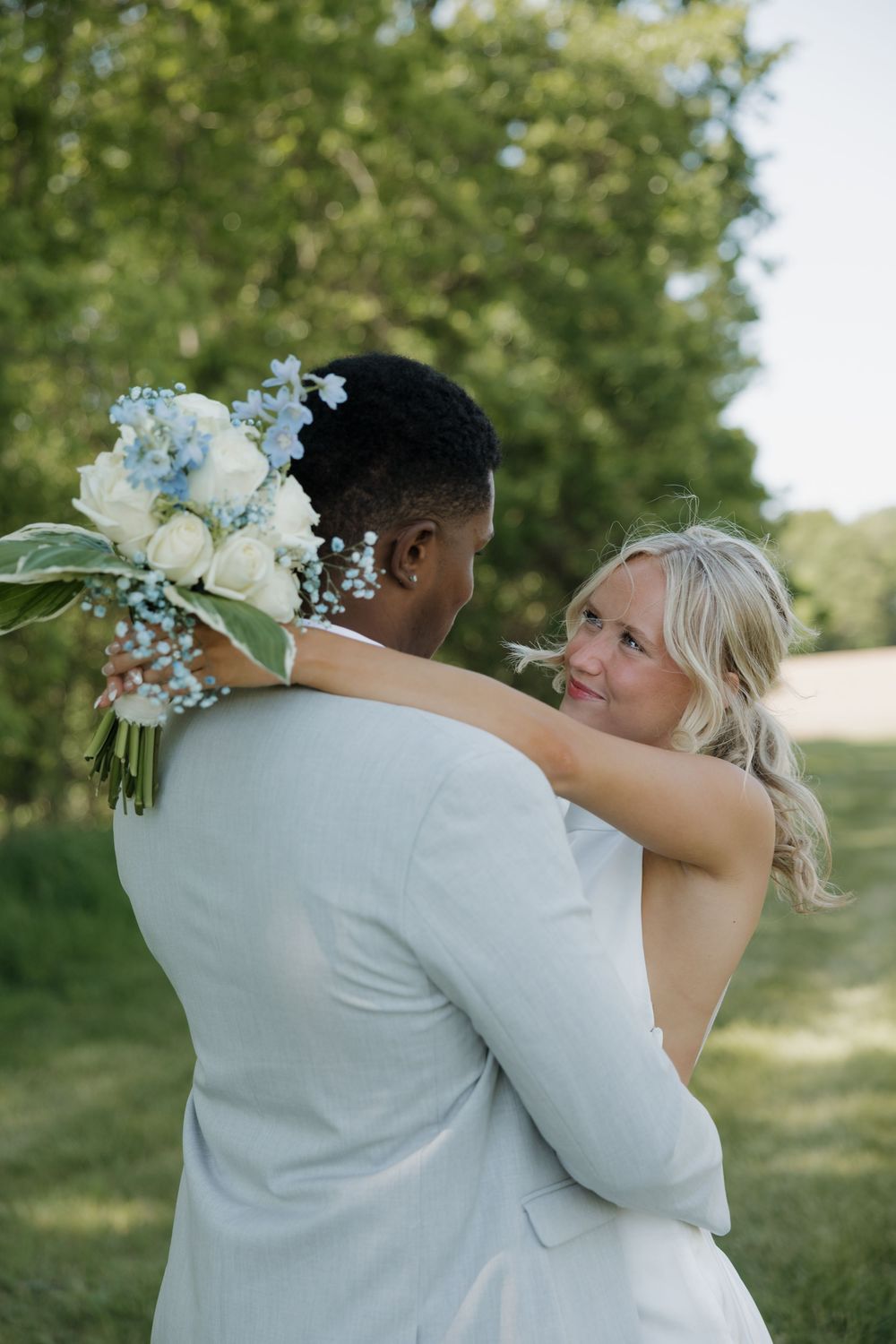 Bride holding blue and white bouquet embraces groom in light suit outdoors at wedding ceremony.