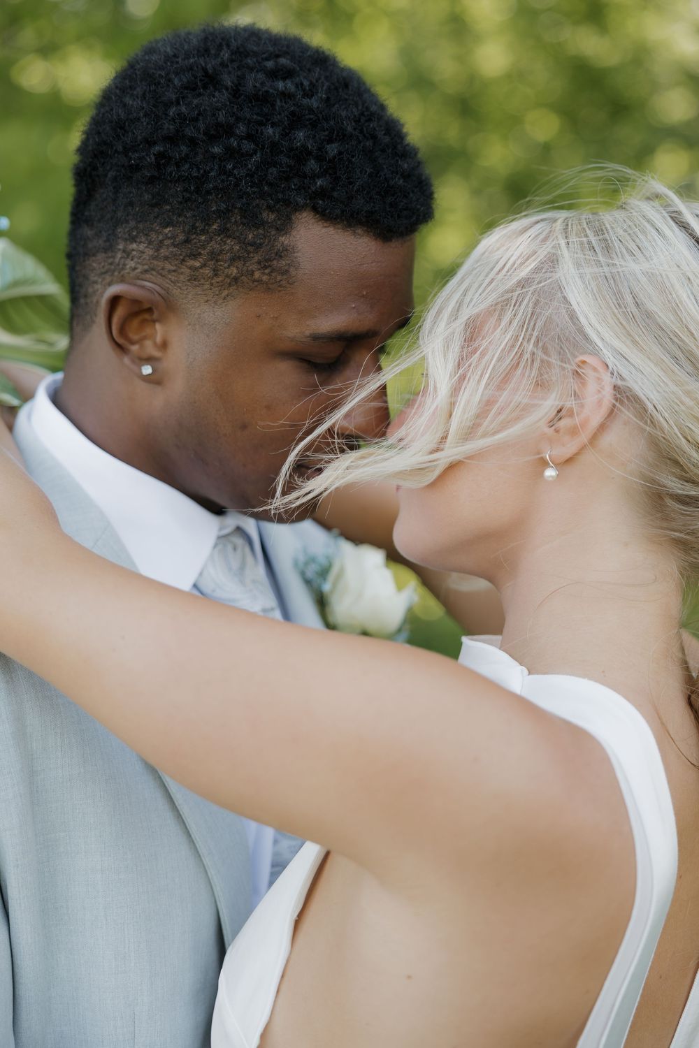Interracial couple sharing a romantic moment at their wedding, groom in gray suit, bride in white dress outdoors.