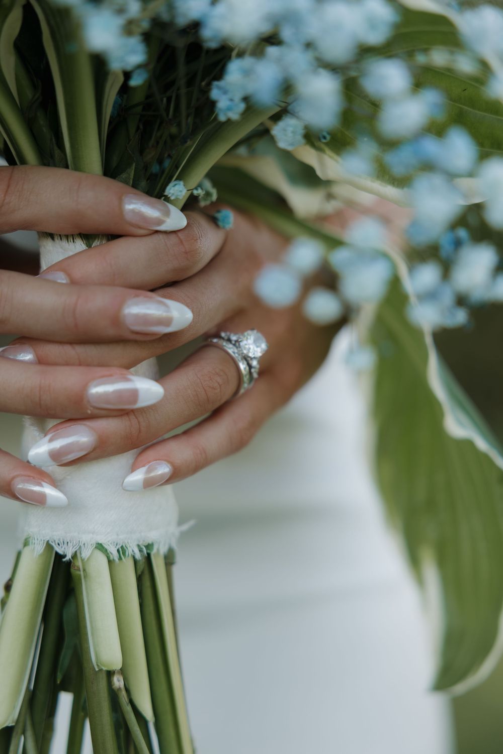Bride holding blue floral bouquet with pearl French manicure nails and diamond engagement ring on wedding day.