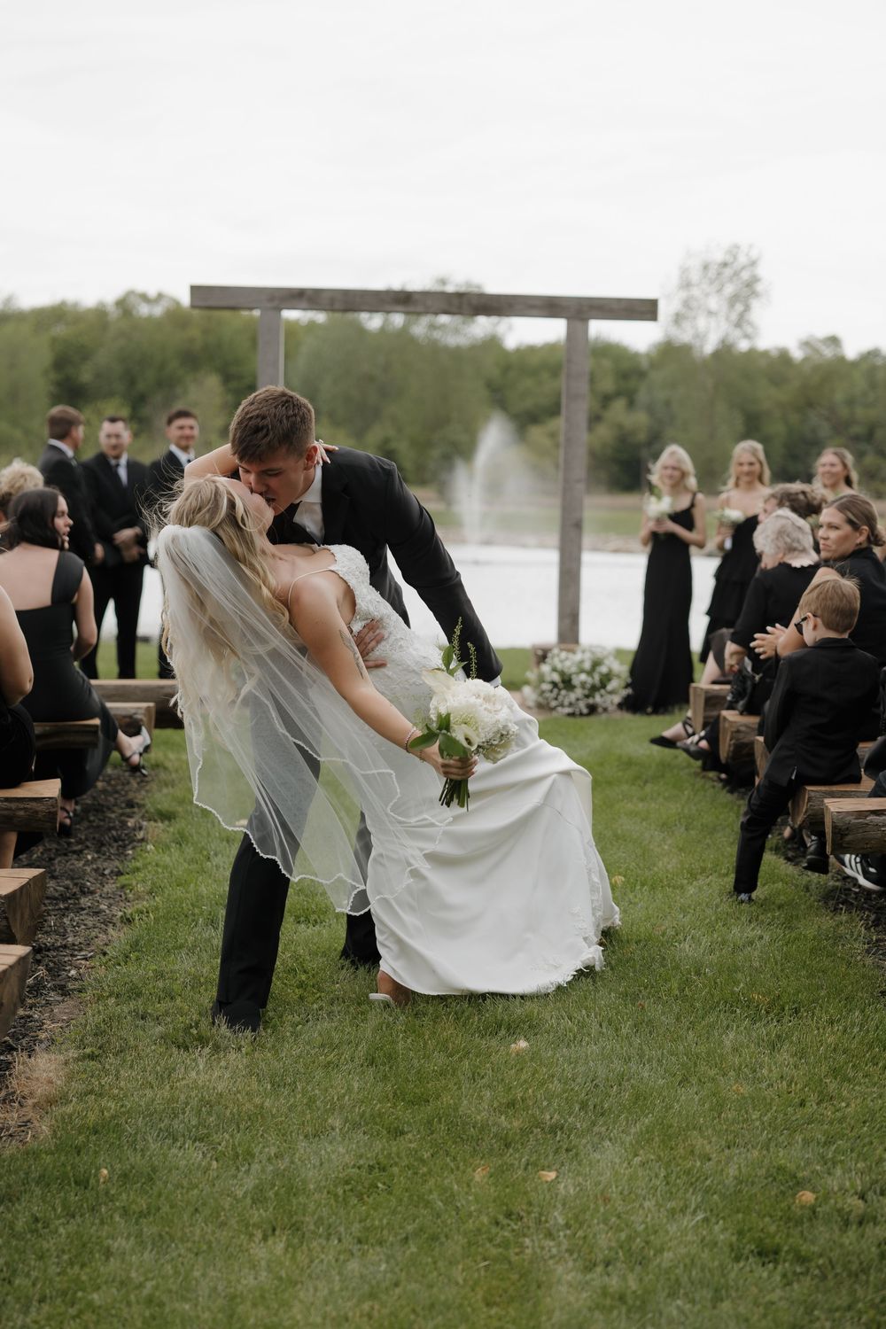 Groom dipping bride for a kiss after outdoor wedding ceremony near a garden fountain.