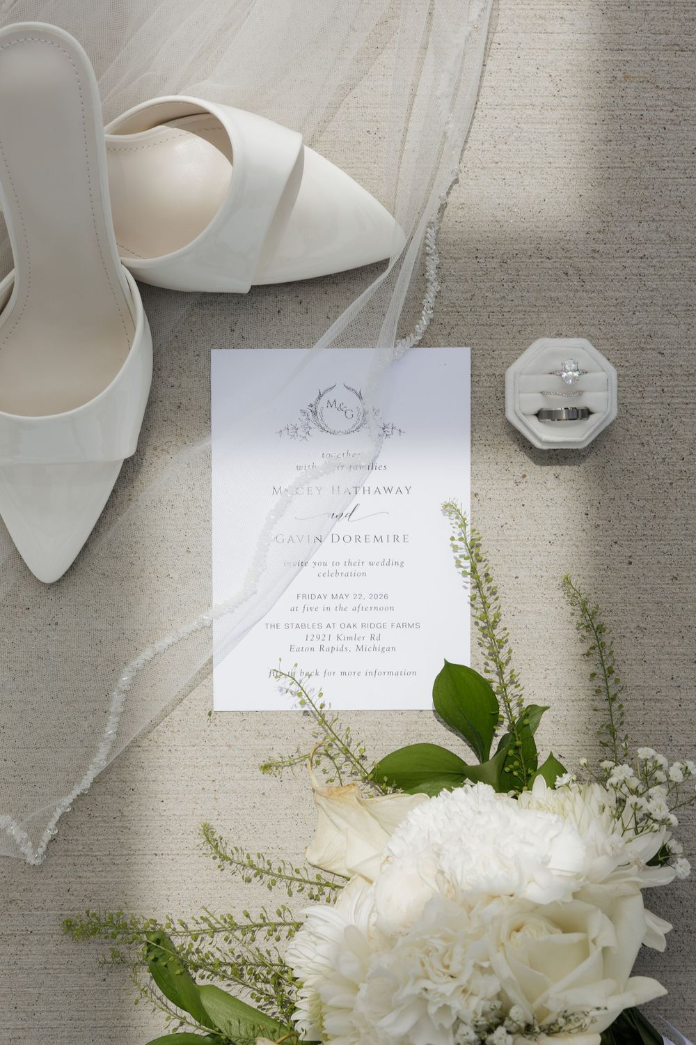 Elegant wedding flat lay with white heels, invitation, diamond ring, and white floral bouquet.
