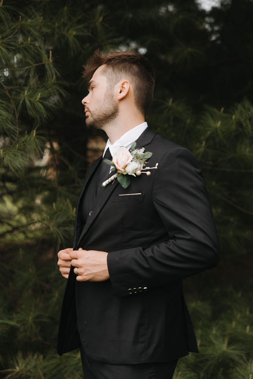 Elegant groom in black suit with floral boutonniere standing near pine trees outdoors.