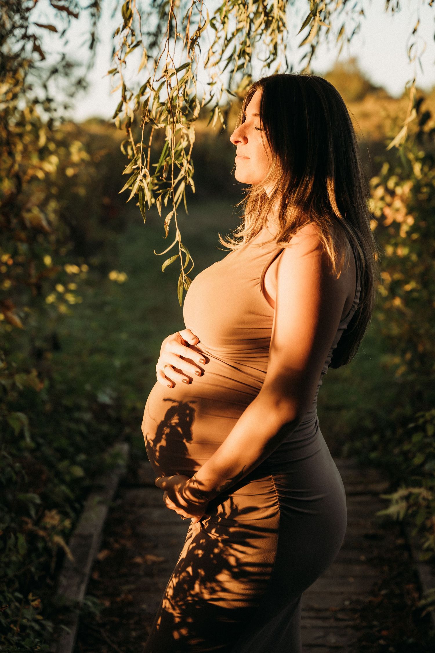 Pregnant woman in beige dress cradling her baby bump outdoors in golden sunset light among lush green trees.