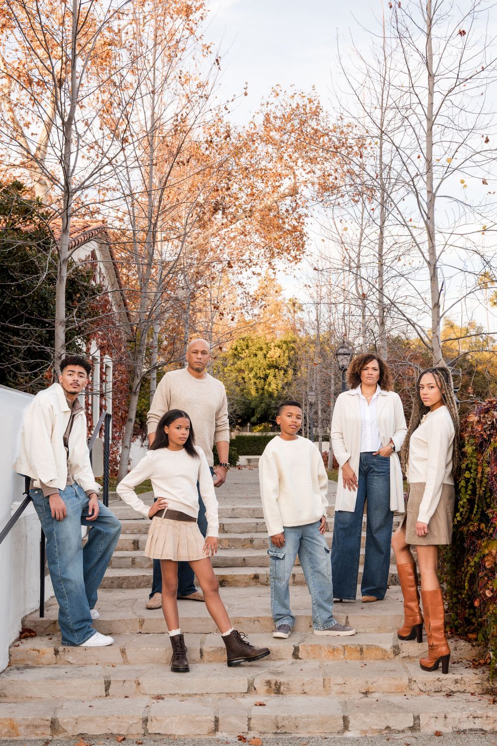 A family of six poses on stone steps surrounded by autumn trees, wearing coordinated cream and neutral outfits.