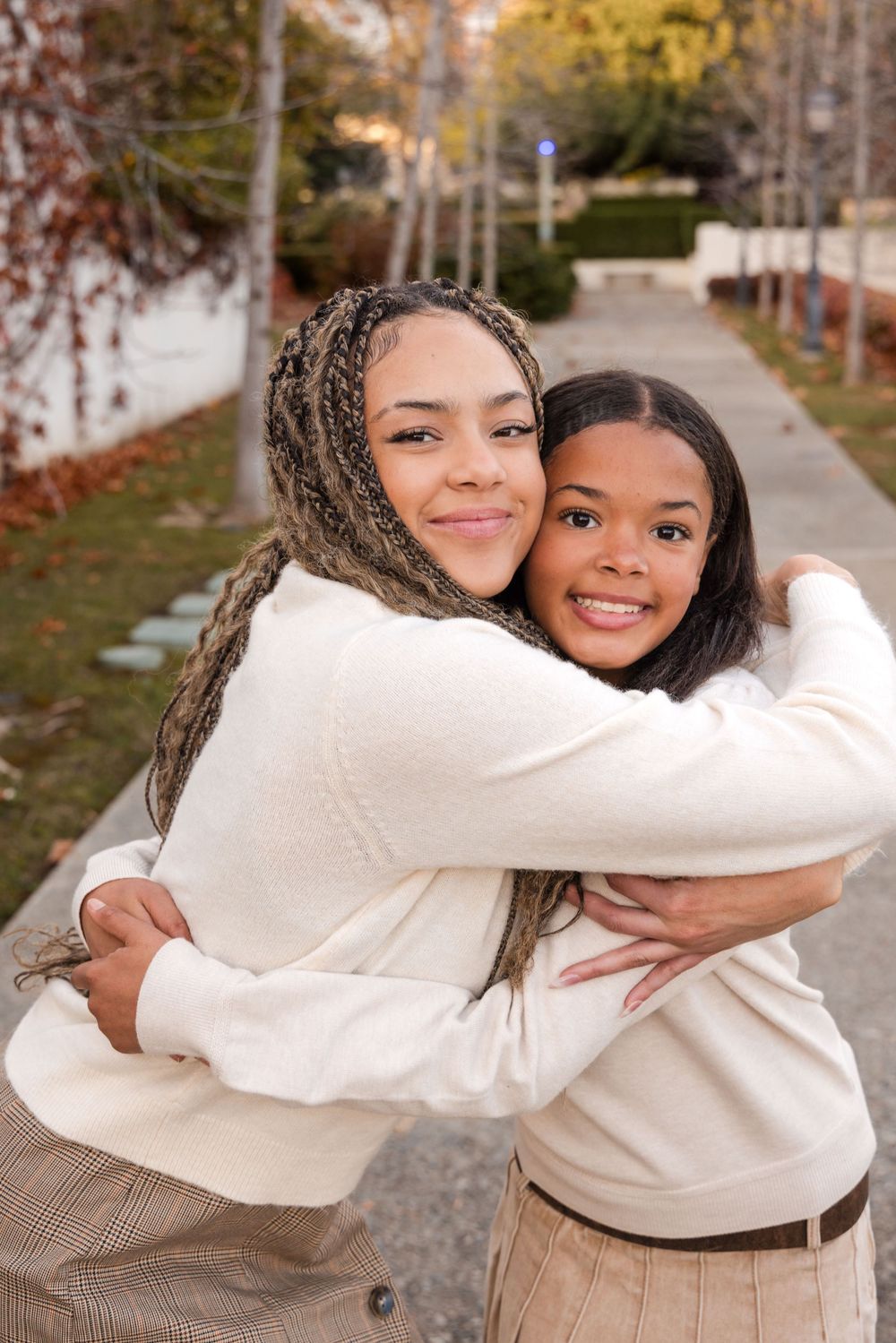 Two young sisters share a warm embrace on an outdoor pathway, smiling at the camera in matching neutral outfits.