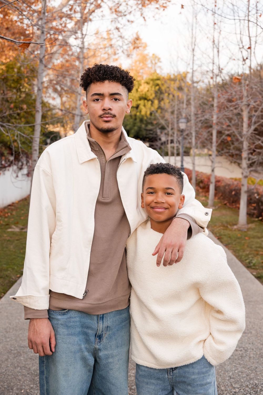 Two brothers pose together outdoors in autumn, with the older teen resting his arm around his younger sibling.