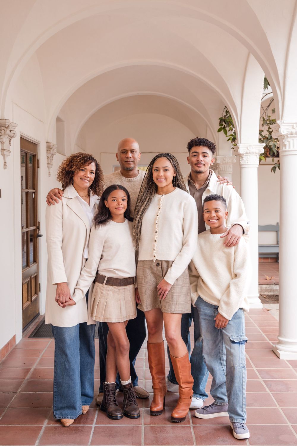A family of six smiles together in a covered archway corridor, wearing coordinated neutral and cream-toned outfits.