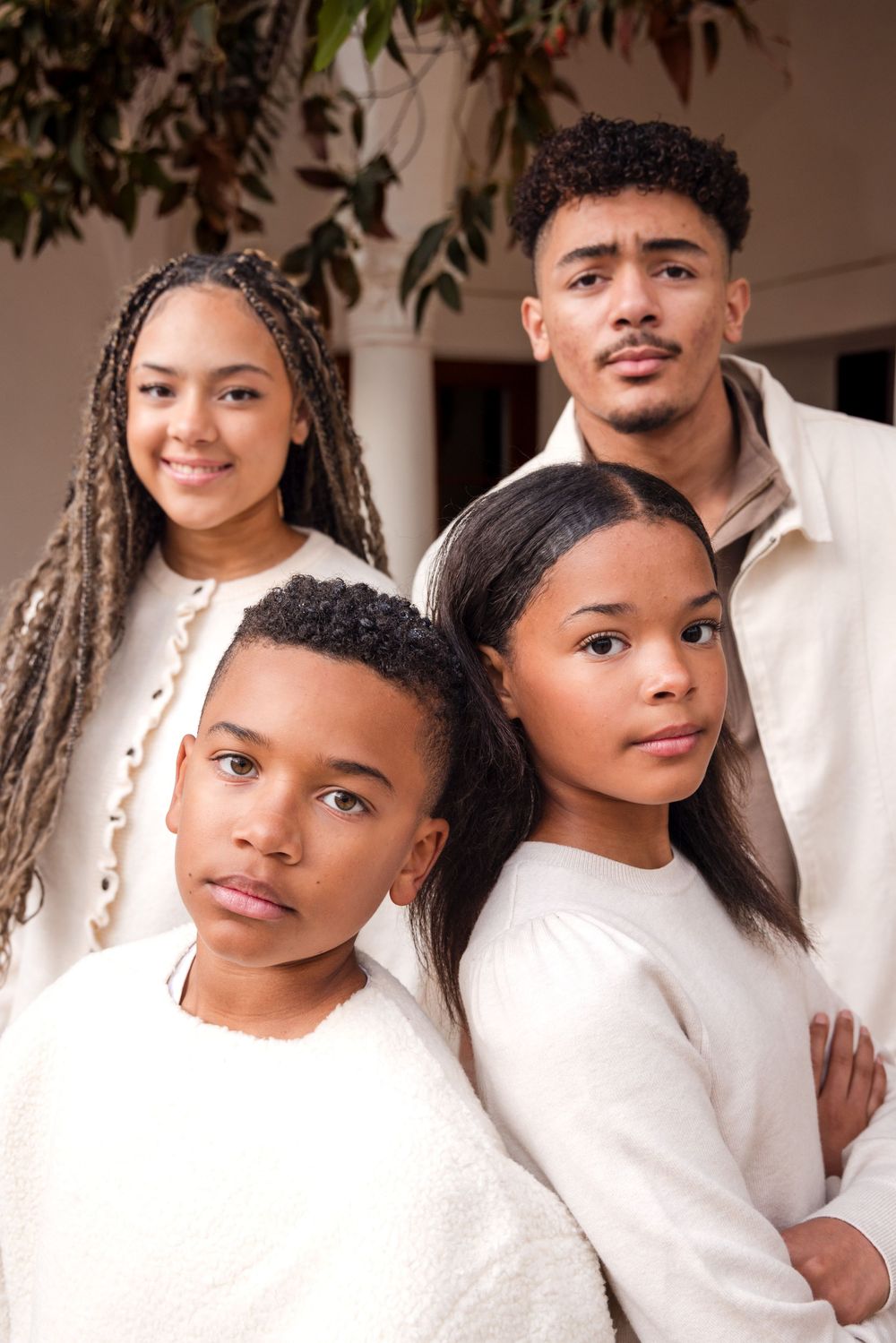 Four siblings pose together for a close-up portrait outdoors, wearing coordinating white and cream-toned outfits.