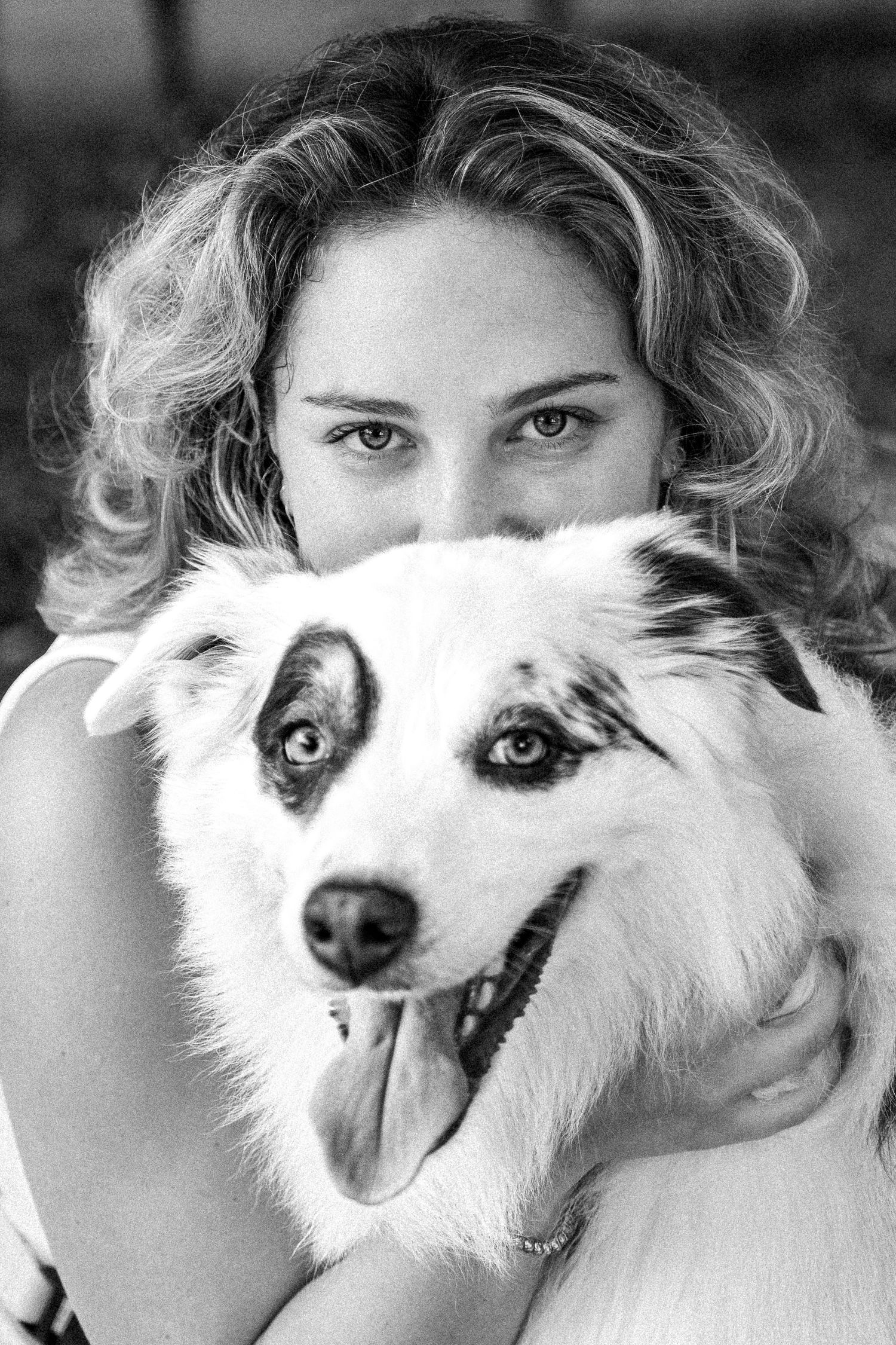 Black and white portrait of a young curly-haired woman hugging a fluffy Border Collie dog with tongue out.