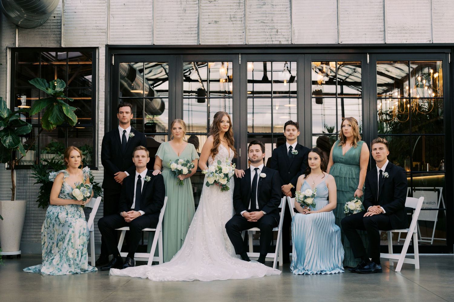 Wedding party poses in front of glass doors, bridesmaids in blue-green gowns and groomsmen in black suits.