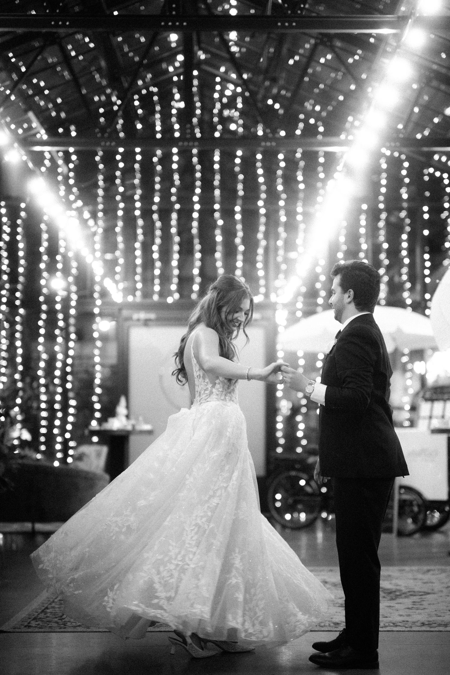 Black and white photo of bride and groom sharing their first dance under twinkling string lights at wedding reception.