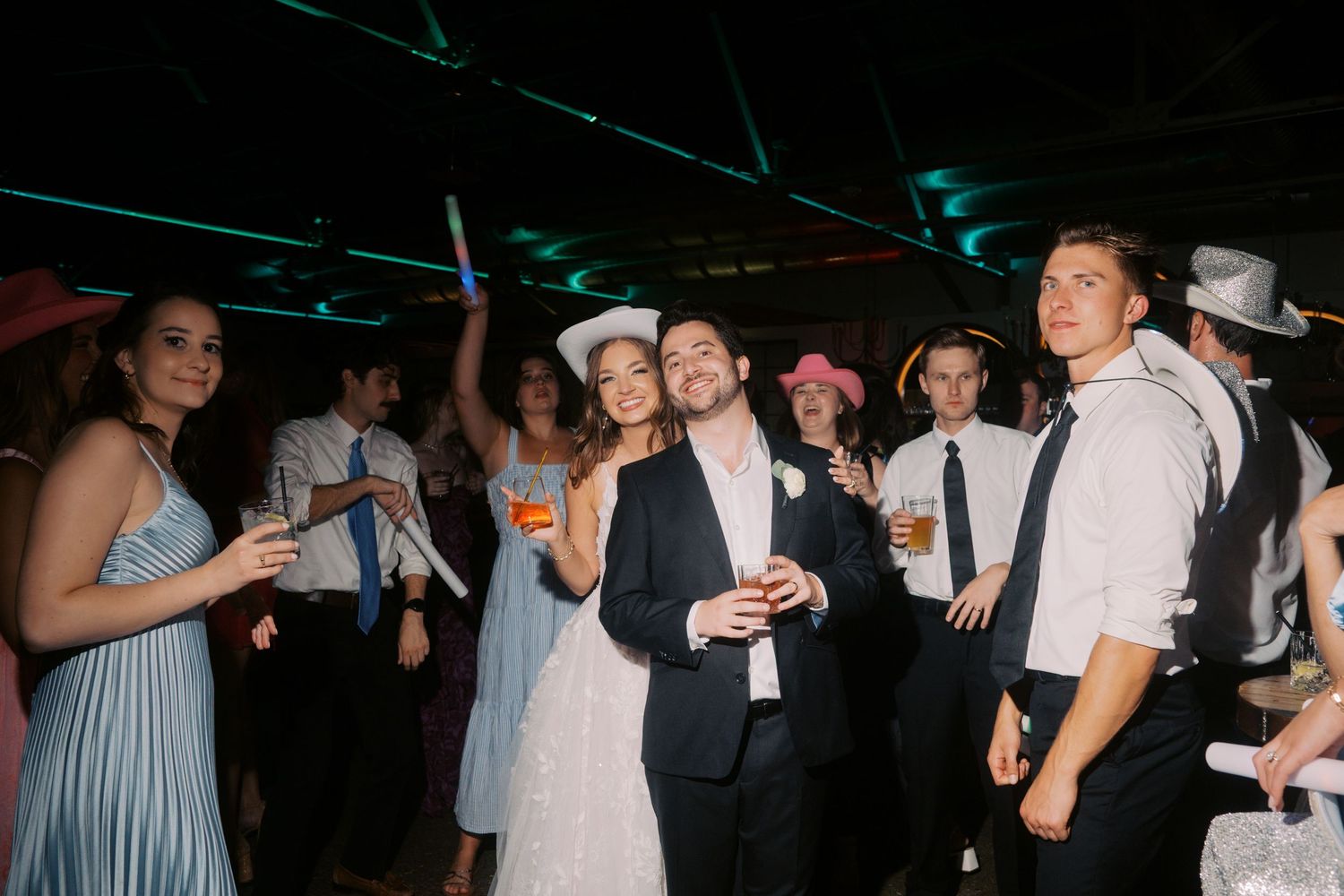 Bride and groom dancing with guests at a lively wedding reception party under colorful lights.