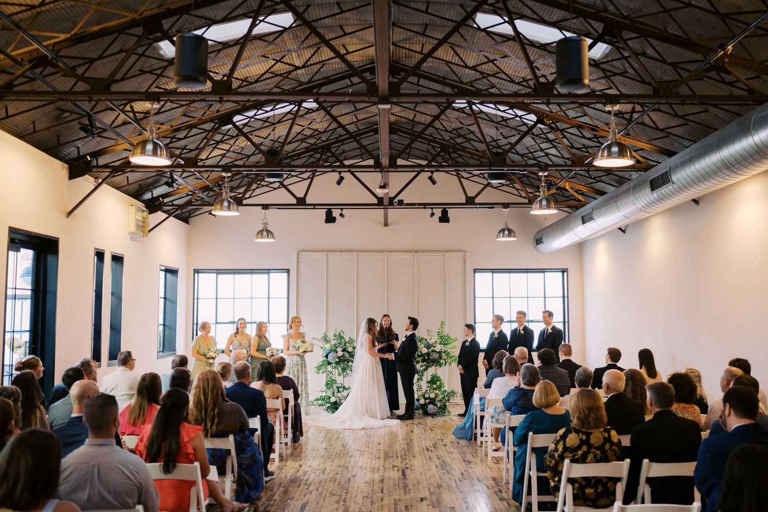 Indoor wedding ceremony in an industrial venue with exposed ceiling, white backdrop, floral arrangements, and seated guests.