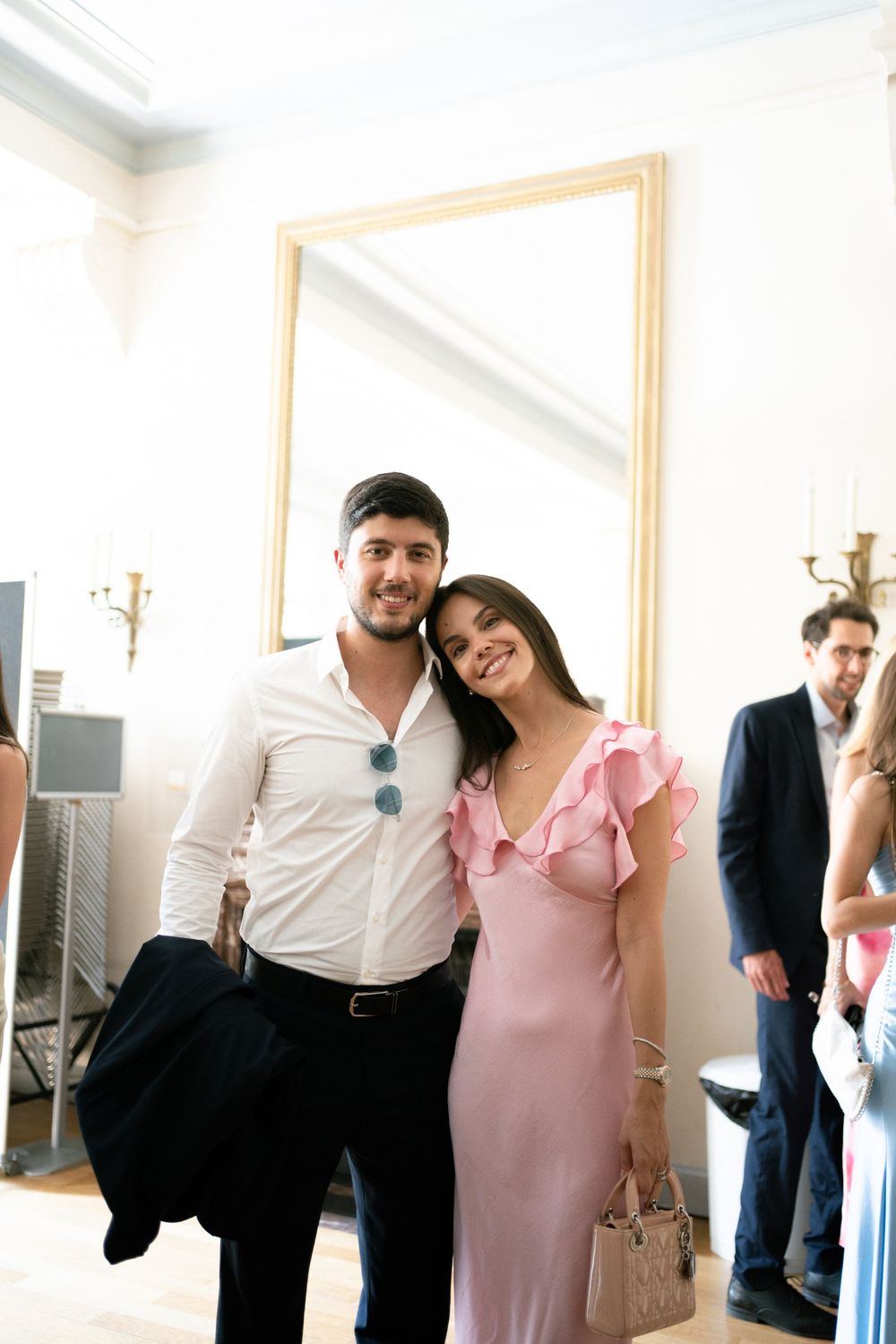 A smiling couple poses together at an elegant event, man in white shirt, woman in pink ruffled dress, large mirror behind them.
