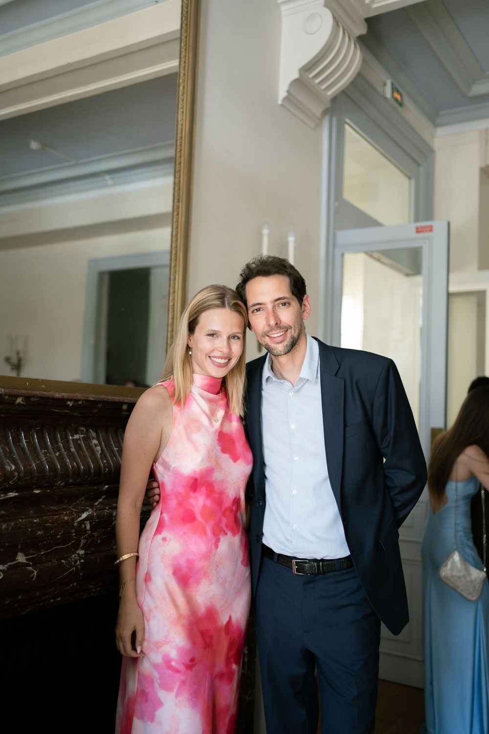A smiling couple poses together at a formal event, woman in pink floral dress, man in navy suit.
